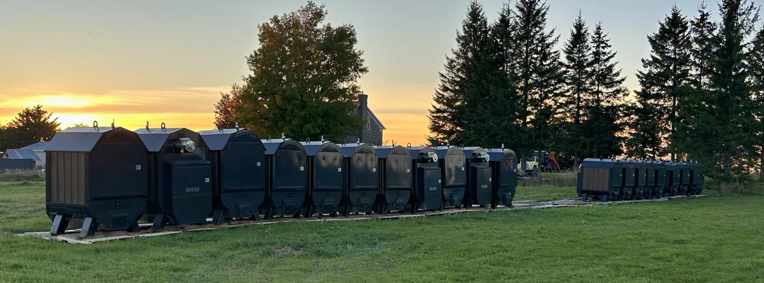 A long row of outdoor wood boilers lined up on pallets in a field at sunset