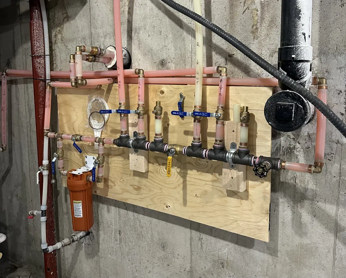 Heating pipework on a basement wall: a black iron manifold with blue and yellow shutoff valves, PEX lines and an orange water filter