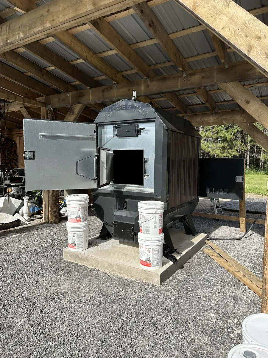 A Portage & Main outdoor wood boiler on a concrete pad under a timber-frame shelter, doors open, with buckets of boiler supplies beside it