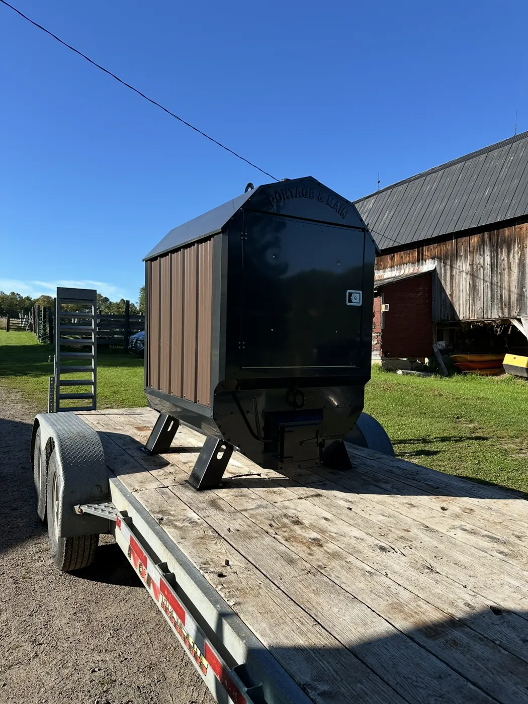 A new Portage & Main outdoor boiler strapped on a flatbed trailer beside a barn